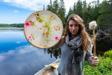 Wall Mural - Beautiful woman holding colorful sacred native frame drum with fur covered drum stick while standing at lake coast Northern Quebec in Canada at day