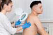 © salomonus_ - A dermatologist examines the skin on the body of a young man through a magnifying glass of a cosmetology lamp. Beauty treatments