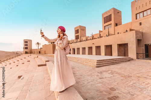 Happy woman traveler wearing dress and turban taking photos on her smartphone of an old Arab town or village in the middle of the desert Canvas Print