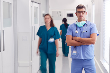 Wall Mural - Portrait Of Mature Male Doctor Wearing Scrubs Standing In Busy Hospital Corridor