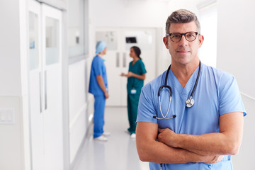 Portrait Of Mature Male Doctor Wearing Scrubs Standing In Busy Hospital Corridor