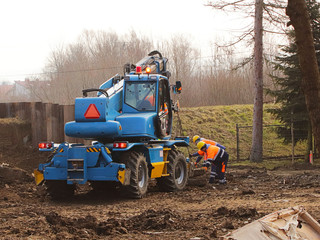 Two workers in bright overalls repair an excavator at the construction site. Landscaping and earthwork. The use of heavy machinery in construction work. Excavated soil under the foundation