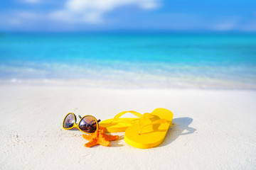 Concept of summer beach holiday. Beach accessories - sunglasses, starfish, yellow flip-flops on sandy tropical beach against blue sky with clouds on bright sunny day. Beautiful colorful image.