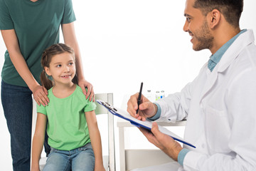 Wall Mural - Smiling pediatrician with clipboard looking at kid near mother isolated on white