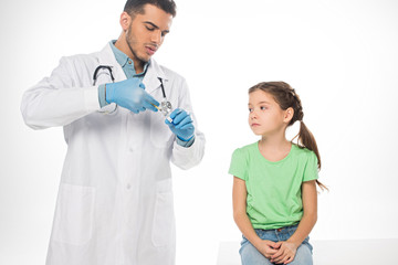 Wall Mural - Handsome pediatrician picking up vaccine in syringe near kid on table isolated on white