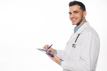 Wall Mural - Side view of smiling doctor with clipboard looking at camera isolated on white