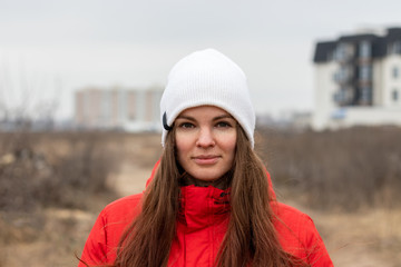 Smiling beautiful young woman with long hair in a white knitted hat and a red jacket with a hood on the street on a cloudy day against a blurred background. Girl without makeup, natural beauty