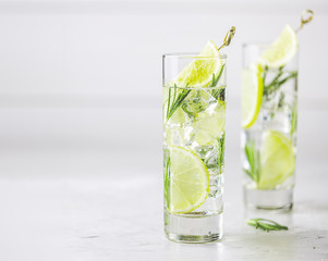 Wall Mural - Refreshing lemon lime drink with ice cubes in glass goblets against a light gray background. Summer fresh lemon soda cocktail with rosemary, selective focus