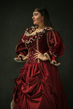 Posing As Royal. Portrait Of Medieval Young Woman In Red Vintage Clothing Standing On Dark Background. Female Model As A Duchess, Royal Person. Concept Of Comparison Of Eras, Modern, Fashion, Beauty.