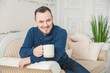 © darkbird - Handsome smiling man with cup of coffee sitting on coach at home