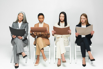 Wall Mural - four multicultural businesswomen in suits sitting on chairs and looking at folders on white