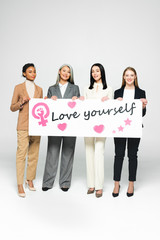 Wall Mural - happy multicultural businesswomen holding poster with love yourself lettering on white