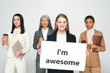 Wall Mural - multicultural businesswomen holding folders and disposable cups near placard with i`m awesome lettering isolated on white
