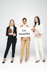 Wall Mural - happy multicultural businesswomen holding folders and paper cups near placard with i`m beautiful lettering on white