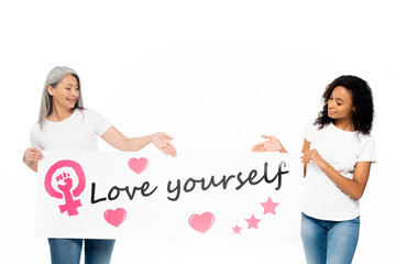 Wall Mural - happy african american and asian women pointing with hands at placard with love yourself lettering isolated on white