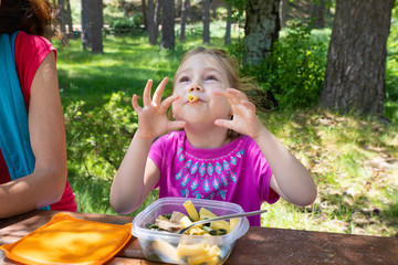 funny four years old blonde girl making fun playing with macaroni, eating pasta from a plastic lunchbox next to her mother in a table picnic in the countryside