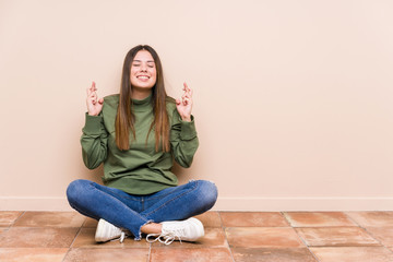 Wall Mural - Young caucasian woman sitting on the floor isolated crossing fingers for having luck