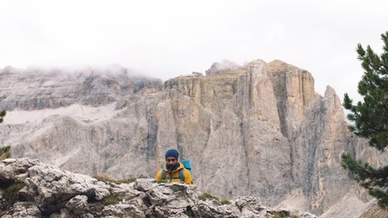 Wall Mural - young man hiker hiking up stone mountain with cloudy sky and fog. yellow jacket, backpack, black bea