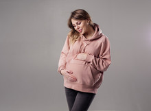 Beautiful Modern Trendy Pregnant Woman Hugs Tummy In Pink Sweatshirt. Gray Photo Studio Background. Pregnancy Expectation Of A Miracle Tenderness Motherly Love. The Future Mom Is Smiling. 9 Months 