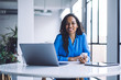 © BullRun - Happy ethnic businesswoman browsing smartphone while working on laptop at office table