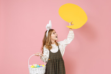 Little pretty blonde kid girl 11-12 years old in spring dress, bunny rabbit ears hold in hand wicker basket colorful eggs Say cloud isolated on pastel pink background. Childhood Happy Easter concept.