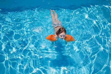 aerial view of four years old blonde child with orange floater sleeves in arms, armbands, swimming in blue transparent water of pool