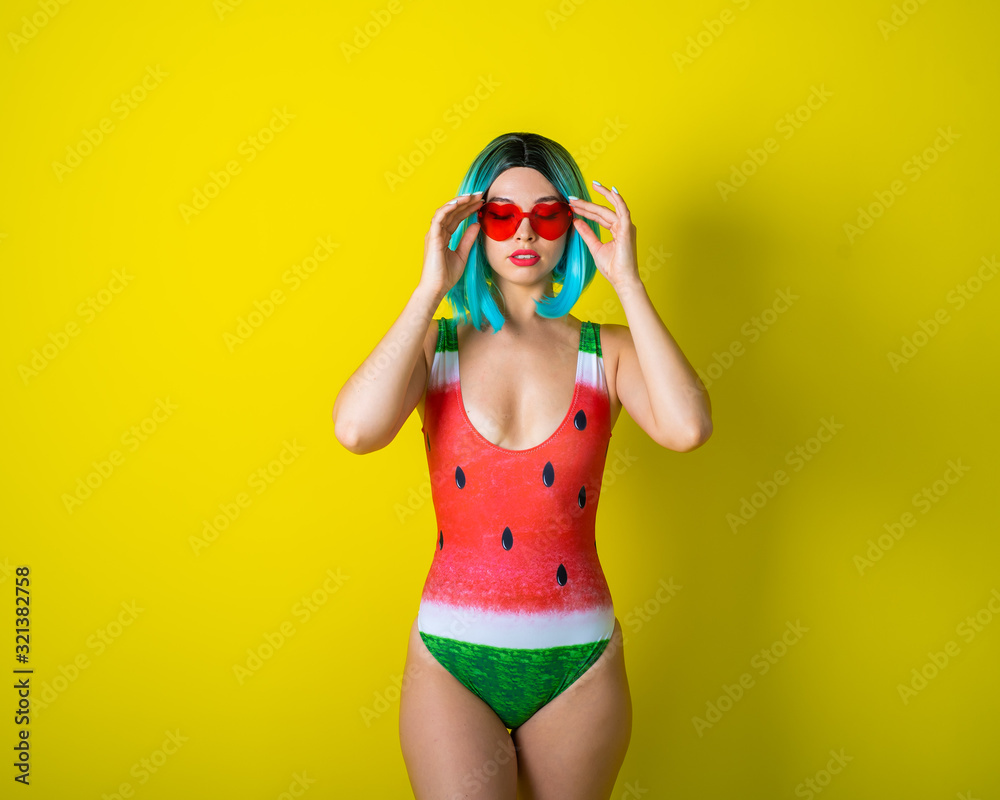 Portrait of a woman in a swimsuit with a picture of a watermelon. Stylish girl in a colored short wig posing in the studio on a yellow background. - obrazy, fototapety, plakaty 