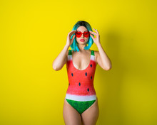 Portrait Of A Woman In A Swimsuit With A Picture Of A Watermelon. Stylish Girl In A Colored Short Wig Posing In The Studio On A Yellow Background.