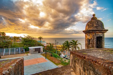 A lookout tower at the Castillo San Cristobal Fort overlooks the colorful houses of La Perla and a basketball court as the setting sun fills the sky with color.