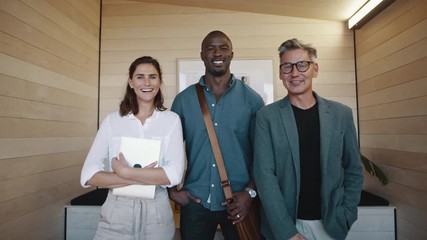 Wall Mural - Three multi-ethnic creatives standing in an office smiling at the camera. Portrait of successful businessmen and businesswoman smiling and looking at camera.