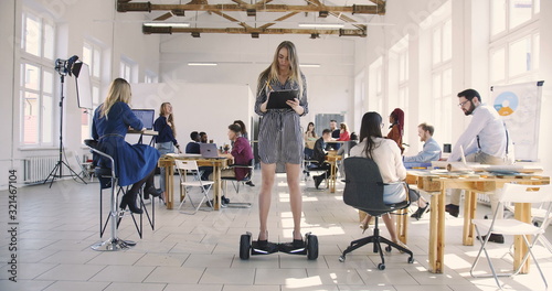 Fotografía Young confident serious blonde business woman in dress riding electric gyroscooter in modern loft multiethnic office