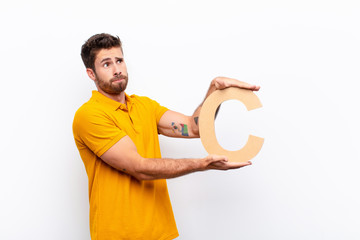 young handsome man confused, doubtful, thinking, holding the letter C of the alphabet to form a word or a sentence.