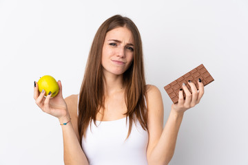 Wall Mural - Young woman over isolated white background having doubts while taking a chocolate tablet in one hand and an apple in the other