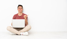 Teenager Man Sitting On The Flor With His Laptop Pointing To The Side To Present A Product
