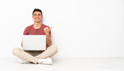 Teenager man sitting on the flor with his laptop pointing to the side to present a product