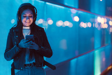 Portrait Of Smiling Teenage Girl With Headphones Standing In Front Of Blue Glass Pane Looking At Smartphone
