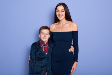 Horizontal shot of cheerful happy mother and son standing isolated over blue background in studio, looking directly at camera, being in good mood. Shoolboy hugging his cute mother in dark blue dress.