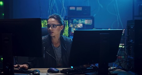 Female beautiful Caucasian developer or hacker in the glasses programming at the two big screen of computers in the dark software room with high technologies.
