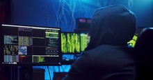 Rear of the close up of the male Caucasian software developer in a hood working and hacking programs or codes at the big screen of computer at night. Back view.