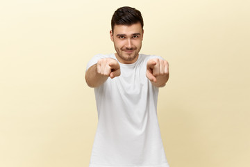 Wall Mural - Horizontal shot of handsome confident positive young guy in white casual t-shirt posing against blank studio wall background with copy space for your content, pointing fore fingers at camera