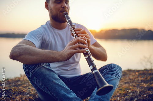 Fotografía Young handsome bearded caucasian blond man sitting on cliff and playing clarinet
