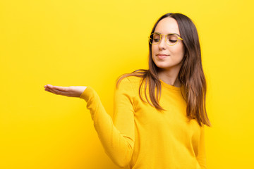 young pretty woman feeling happy and smiling casually, looking to an object or concept held on the hand on the side against orange wall