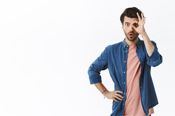 Wall Mural - Curious good-looking handsome bearded man in hipster outfit, showing okay, ok or confirmation sign over eye, folding lips say wow, staring wondered and impressed, white background