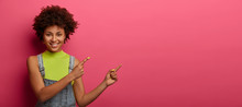 Studio Shot Of Smiling Curly Woman Wears Bright Vest And Dungarees, Points Both Index Fingers At Upper Right Corner, Demonstrates Good Copy Space For Your Promotional Content, Isolated On Pink Wall