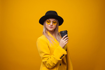 Wall Mural - Take-away coffee. Stylish trendy young woman in bright clothes on a yellow background.