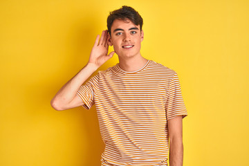 Wall Mural - Teenager boy wearing yellow t-shirt over isolated background smiling with hand over ear listening an hearing to rumor or gossip. Deafness concept.