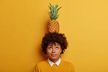 Portrait Of Curly Haired Ethnic Woman Stands With Tropical Fruit On Head, Looks Above, Shows How She Can Carry Pineapple, Wears Yellow Sweater, Stands Against Vibrant Background. Monochrome.