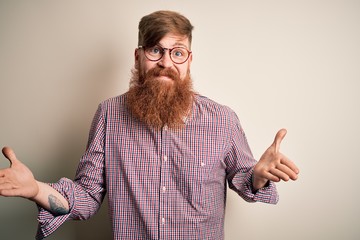 Wall Mural - Handsome Irish redhead business man with beard wearing glasses over isolated background clueless and confused expression with arms and hands raised. Doubt concept.