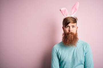 Canvas Print - Hipster Irish man with beard wearing easter rabbit ears over isolated pink background with serious expression on face. Simple and natural looking at the camera.