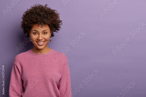Fotografía Portrait of lovely glad woman has Afro hair, smiles joyfully, gets pleasant news, gazes at camera cheerful, expresses upbeat emotions, models against purple background